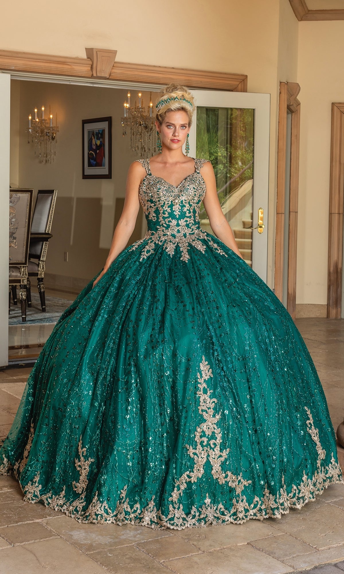 A woman stands indoors wearing the Dancing Queen Quinceanera Dress 1478, a green ball gown featuring gold embroidery, embellished cap sleeves, and a lace corset bodice. She poses near open doors with chandeliers and framed art in the background.